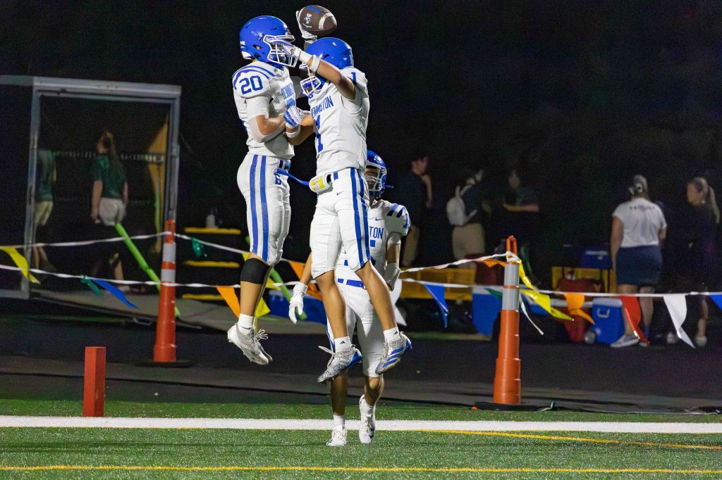 Two football players celebrating a touchdown by jumping together in the air at night, with colorful flags and onlookers in the background.