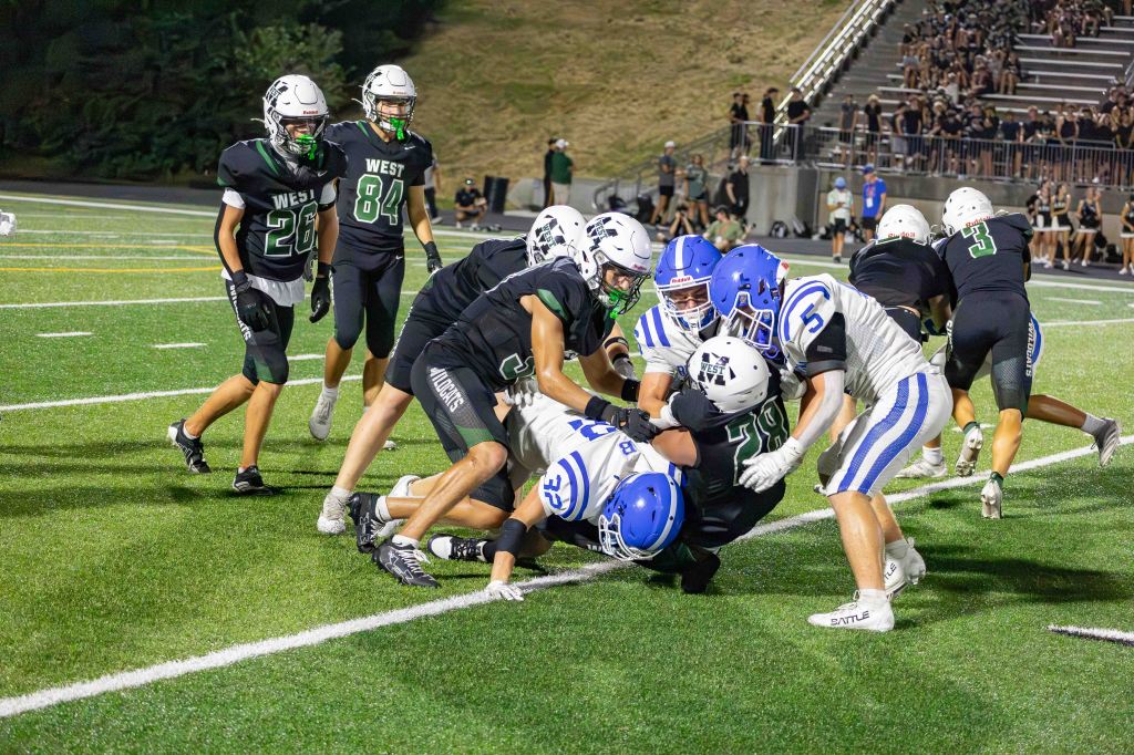 High school football players in action, with several players from both teams engaged in a tackle on the field under stadium lights.