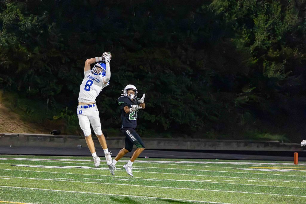 A high school football player in a white jersey with the number 8 catches a pass while being defended by an opponent in a black jersey during a night game.