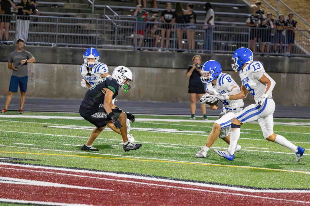 A high school football game in progress with a player in a black uniform being pursued by two players in white uniforms. The background features spectators in an outdoor stadium.