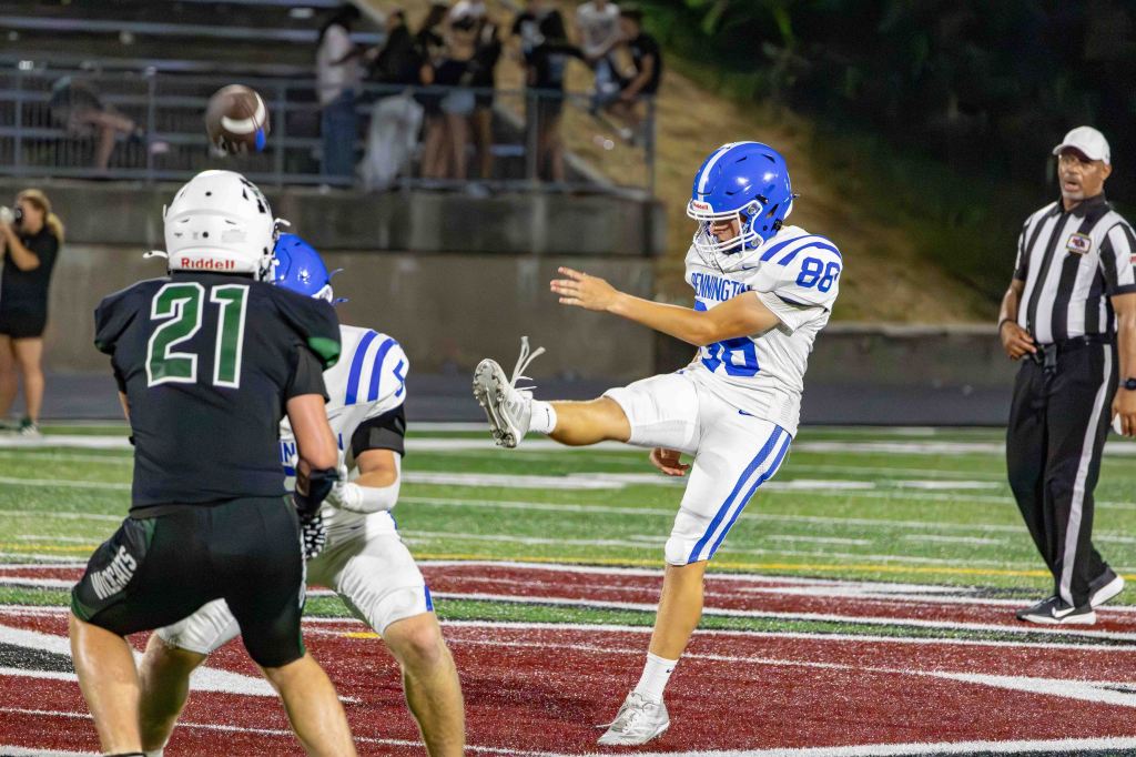 A football player wearing a blue helmet and white uniform with the number 86 kicks a football on a field, while another player in a black uniform with the number 21 is positioned to receive the ball.