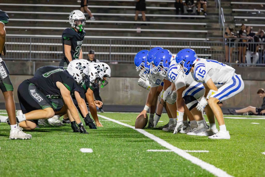 A high school football game scene showing two teams lined up at the line of scrimmage. One team in black uniforms with green accents and the other in blue and white uniforms. Players are preparing for the snap on a well-maintained artificial turf field, with spectators in the background.