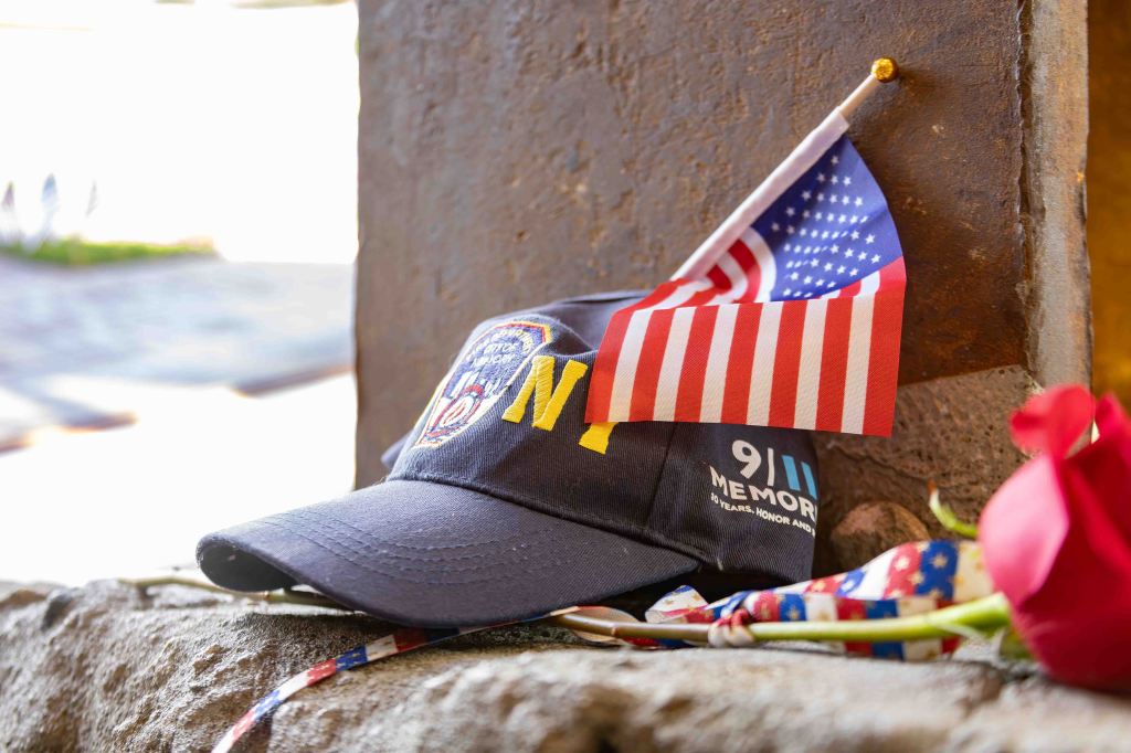 A navy blue cap with 'NY' and a firefighter emblem, placed on a stone surface with a small American flag and a red rose nearby, symbolizing remembrance.
