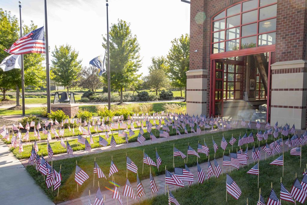 A landscaped area in front of a building with several small American flags displayed in neat rows on the grass, alongside tall trees and flagpoles.