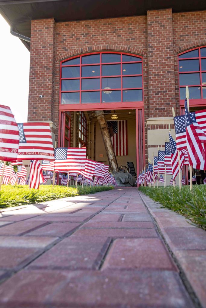 A pathway lined with small American flags leading to a brick building, with large windows and an entrance displaying more flags inside.