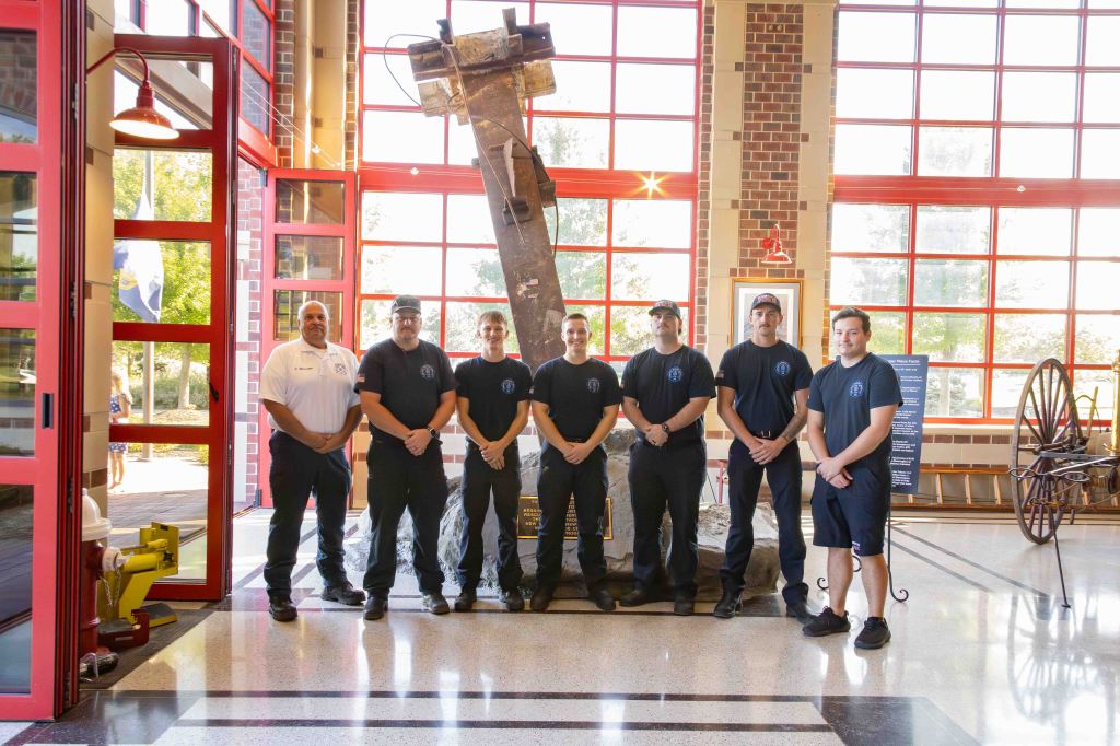 A group of six firefighters in uniform, standing inside a fire station with large red windows, alongside a historical fire engine and equipment display.