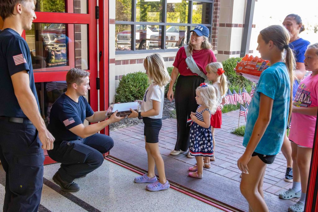A firefighter hands a small package to a girl while a group of children and adults watch. The scene takes place at a fire station with red doors and visible outdoor greenery.