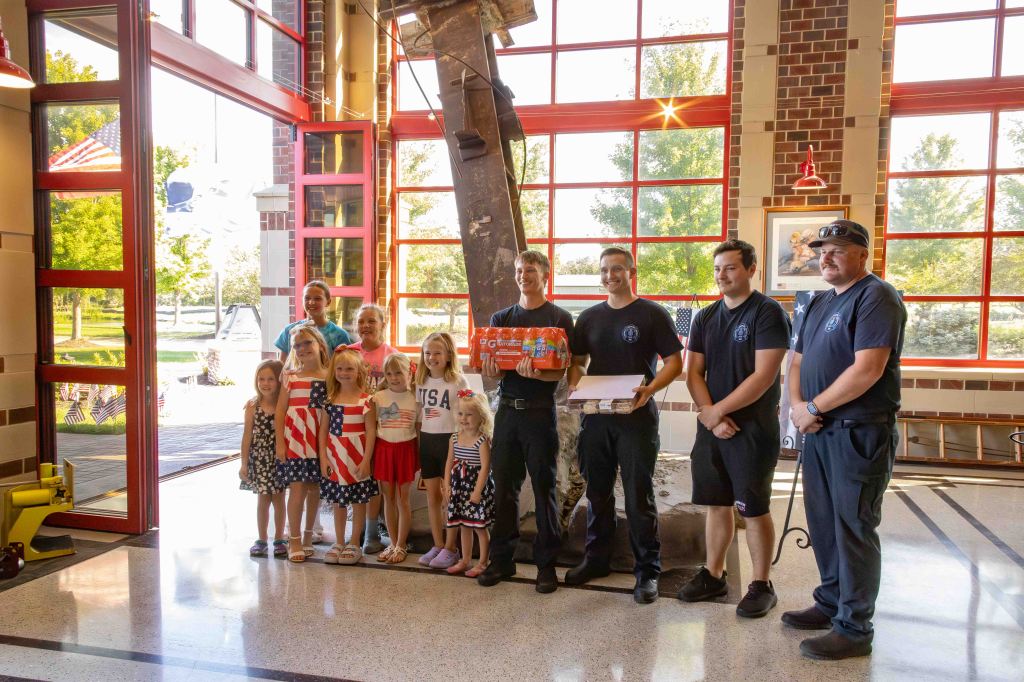 A group of children in patriotic outfits stands in front of firefighters at a fire station, holding gifts and posing for a photo.