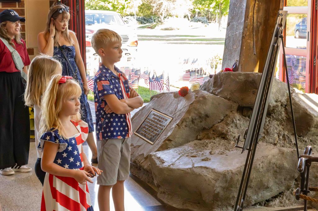 A group of children and adults observing a memorial display with a rock, plaque, and floral tributes, set against a backdrop of American flags.