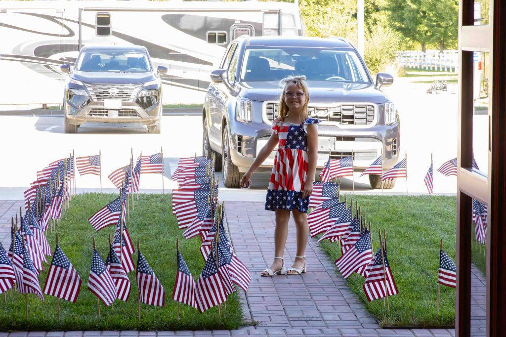 A young girl in an American flag dress stands among rows of small American flags on a grassy lawn, with vehicles parked in the background.