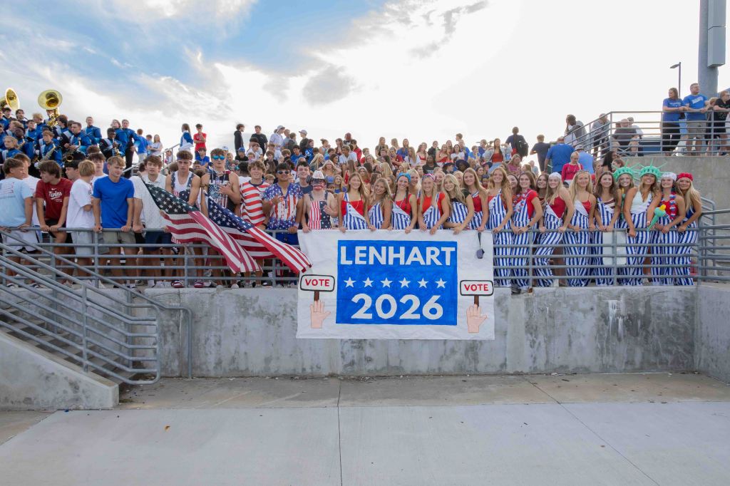 A large group of students dressed in patriotic outfits, holding American flags and a banner that reads 'LENHART 2026' with a 'VOTE' symbol, gathered together at a school event.