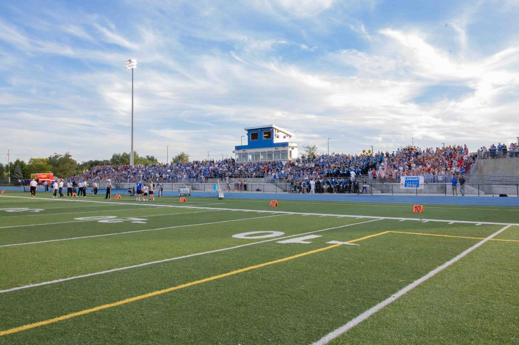 A wide view of a football field filled with players and officials, with a large crowd of spectators seated in the bleachers in the background, under a cloudy sky.