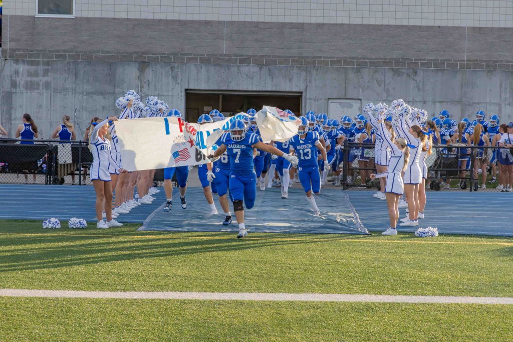 High school football team running onto the field through a banner held by cheerleaders, with fans cheering in the background.