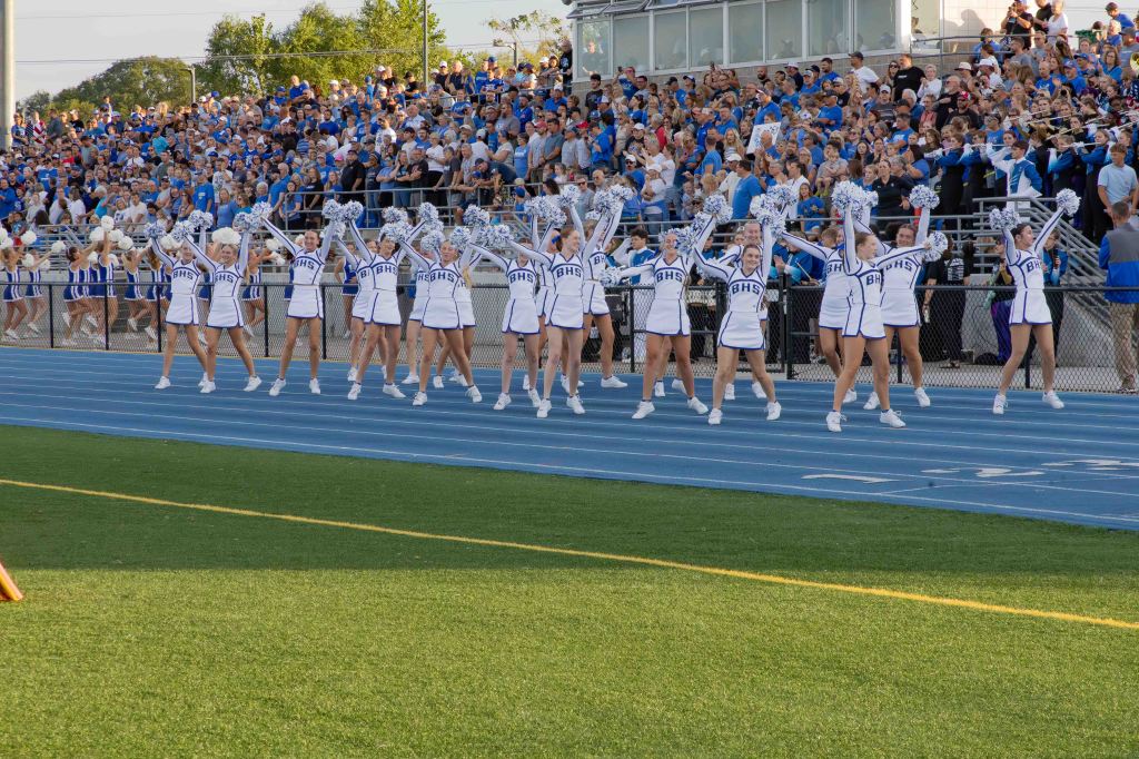 A group of cheerleaders in white and blue uniforms performing together on a track in front of a large crowd at a sports event.
