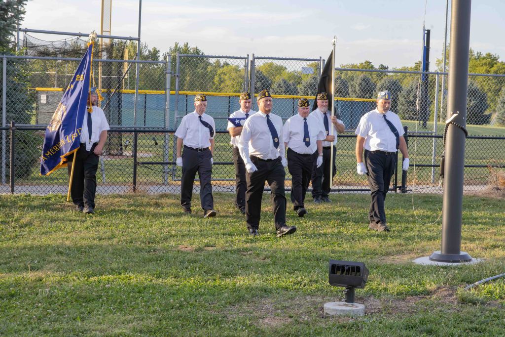 A group of several men in uniform, including a flag bearer, walking in formation on a grassy area near a sports field.