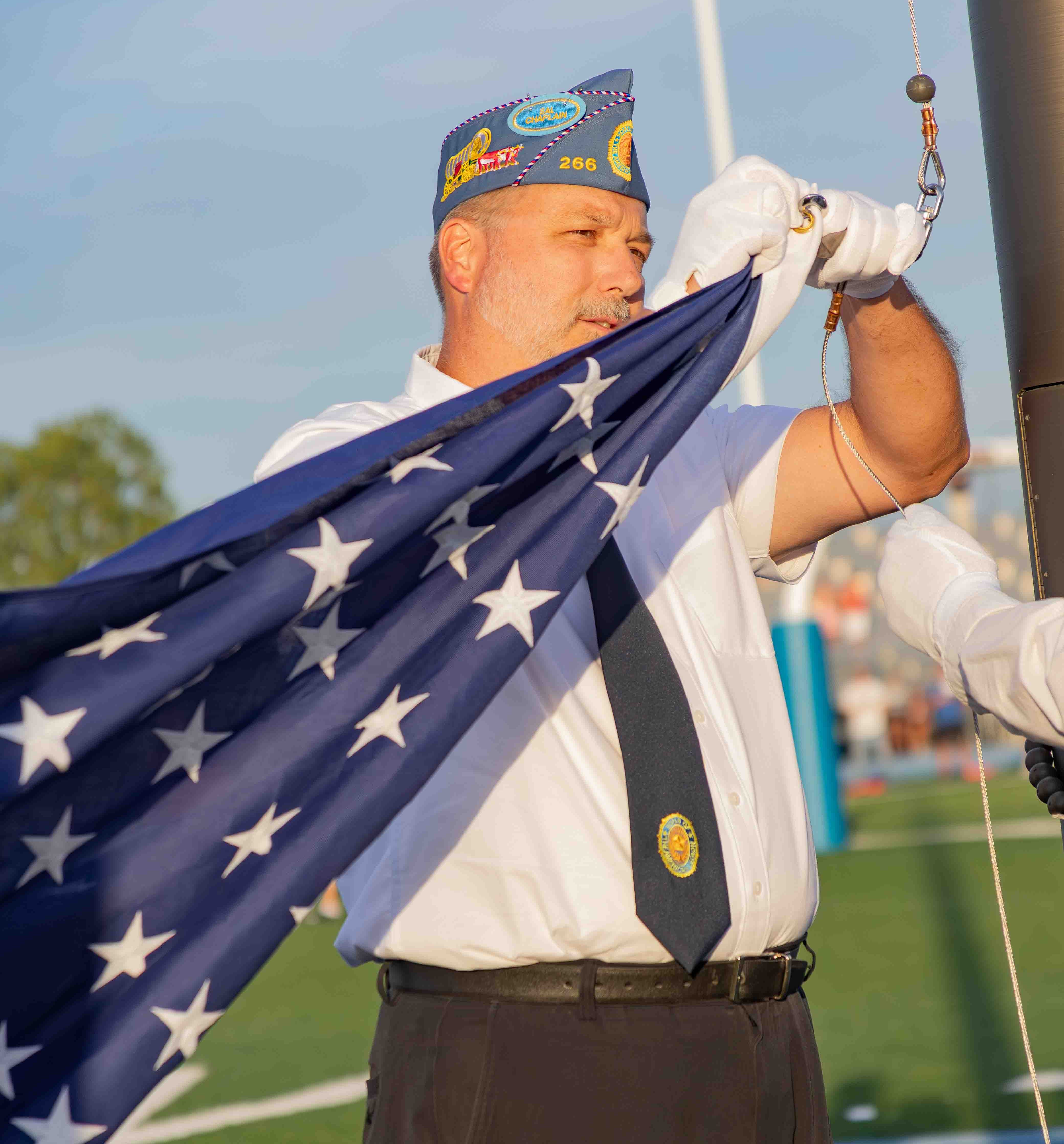 A man in a military-style cap and white shirt is preparing to raise an American flag, holding it with one hand while adjusting the mechanism with the other.