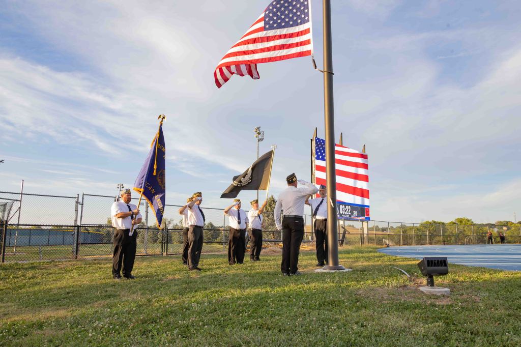 Veterans standing in formation during a flag ceremony, with American flags displayed and a scoreboard in the background.