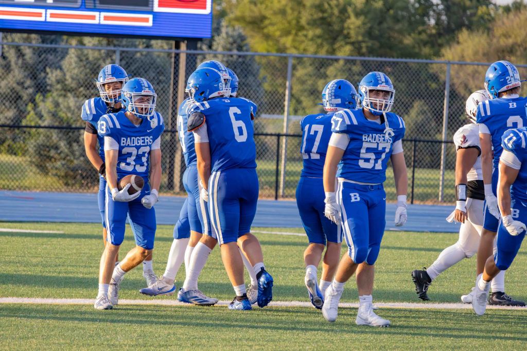 A group of high school football players wearing blue uniforms with 'Badgers' on them, celebrating on the field. The players are engaged and focused as they prepare for the next play.