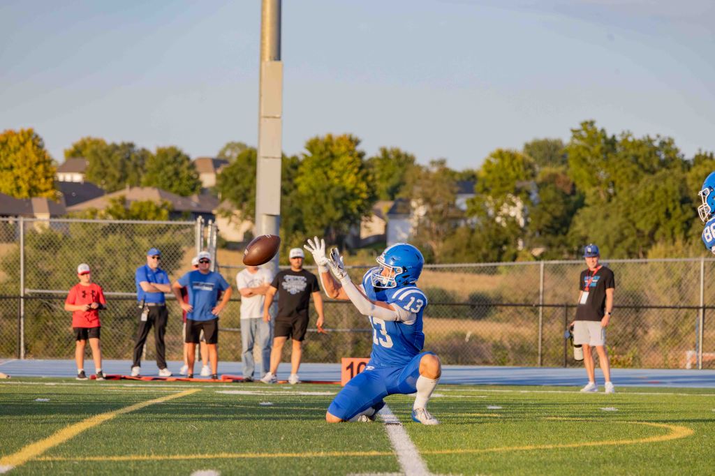 A football player in blue uniform, number 13, kneels on the field reaching for a football while teammates and spectators watch in the background.