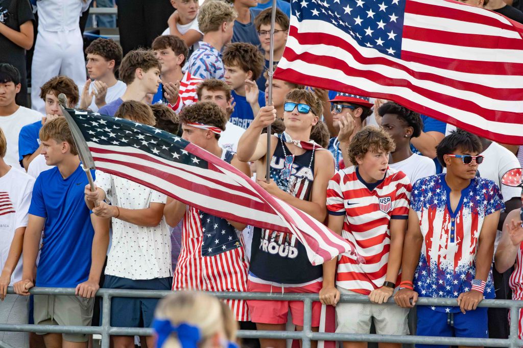 Group of young fans at a sporting event, wearing patriotic clothing and holding American flags, cheering and celebrating.