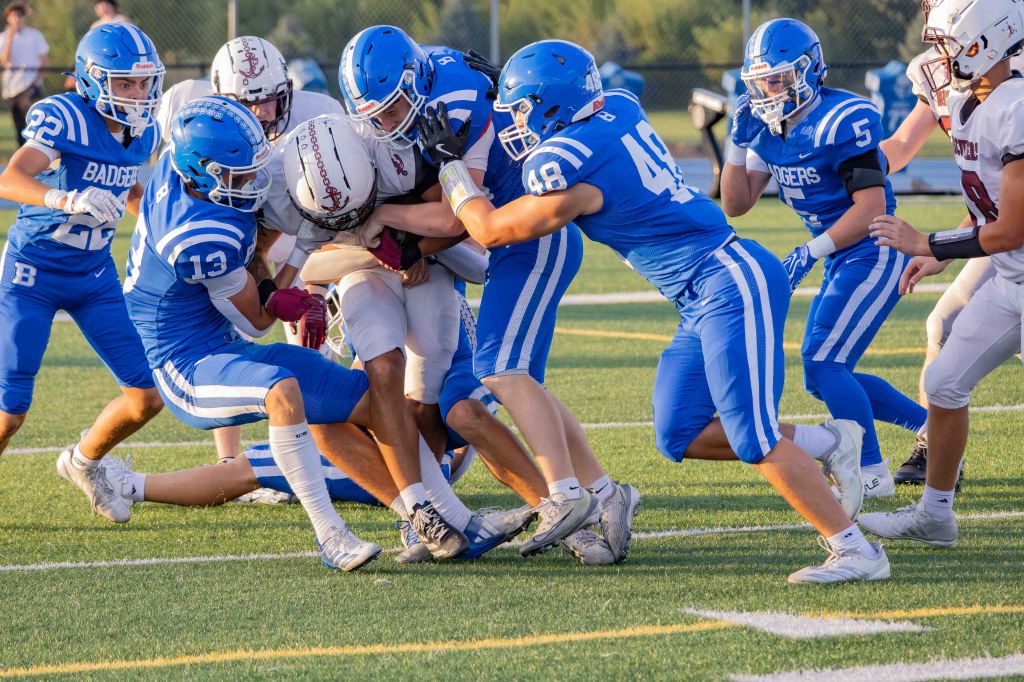 A high school football game in progress, featuring players in blue and white uniforms tackling an opposing player in a white jersey with a red helmet.