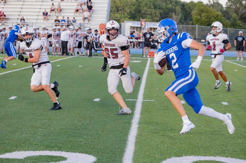 A football player in a blue uniform running with the ball on the field, with two defenders in white uniforms chasing from behind.