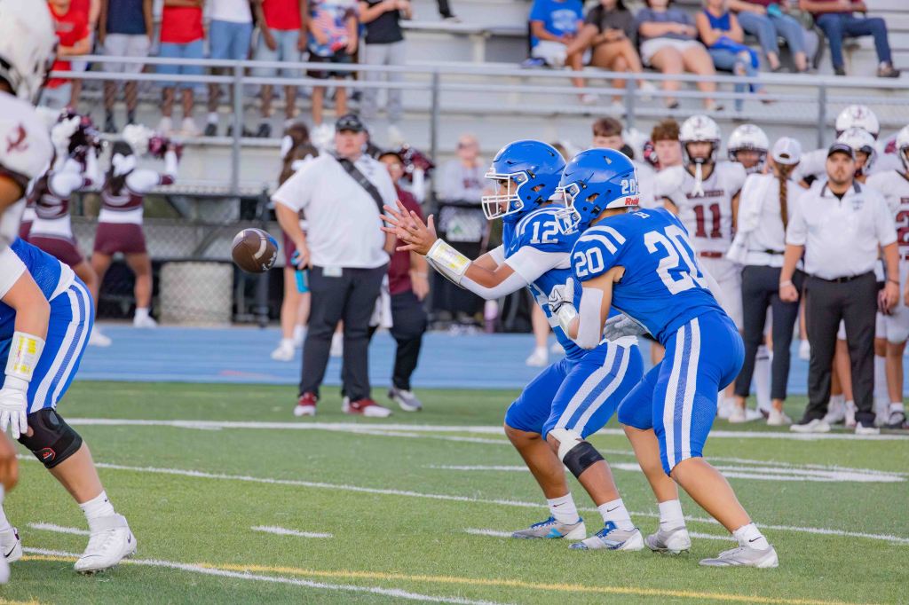 A youth football game in progress, featuring two players in blue uniforms, one preparing to receive a snap from the center. In the background, spectators and cheerleaders are visible.