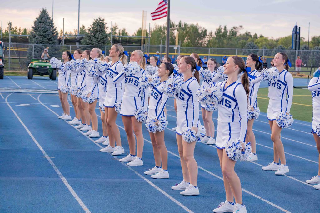 A group of cheerleaders in white and blue uniforms, standing on a blue track, performing a cheer routine. They are holding pom-poms and smiling, with an American flag visible in the background.