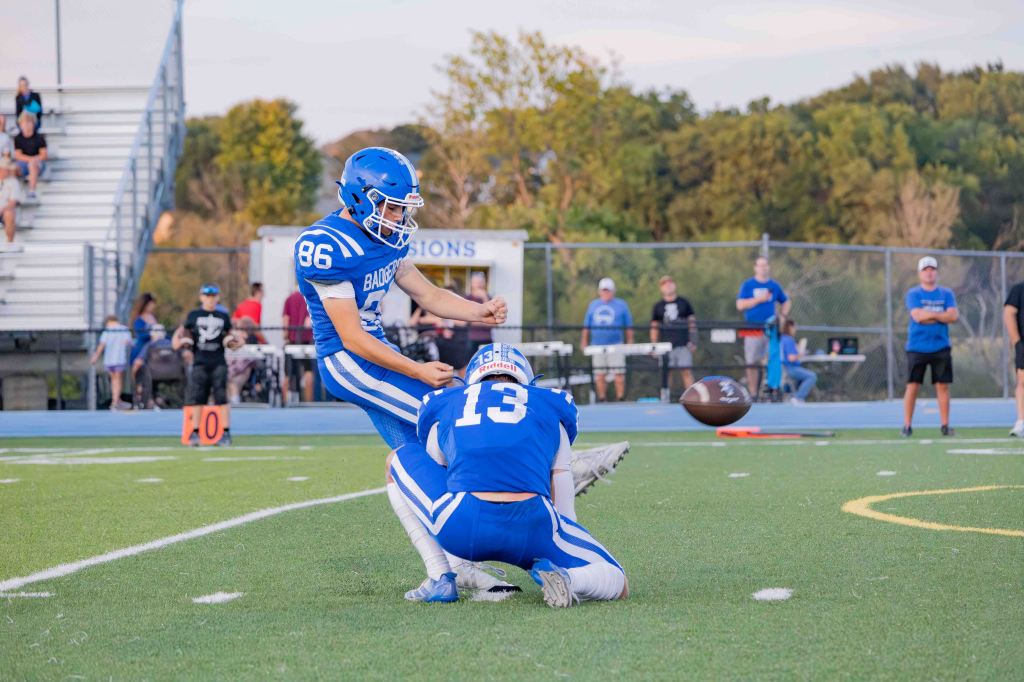A football player wearing number 86 prepares to kick the ball while another player in blue, number 13, holds it on the ground during a game. Spectators are visible in the background.