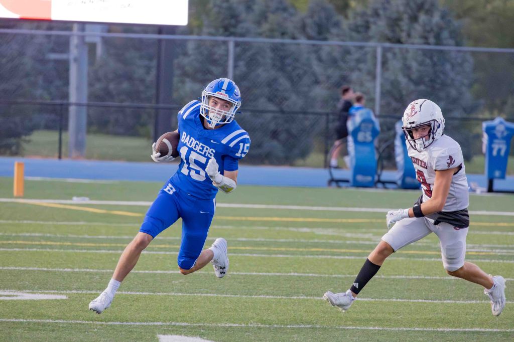 A high school football player in a blue jersey with 'Badgers' printed on it runs with the ball, while a player in a white jersey chases him on a grassy field.