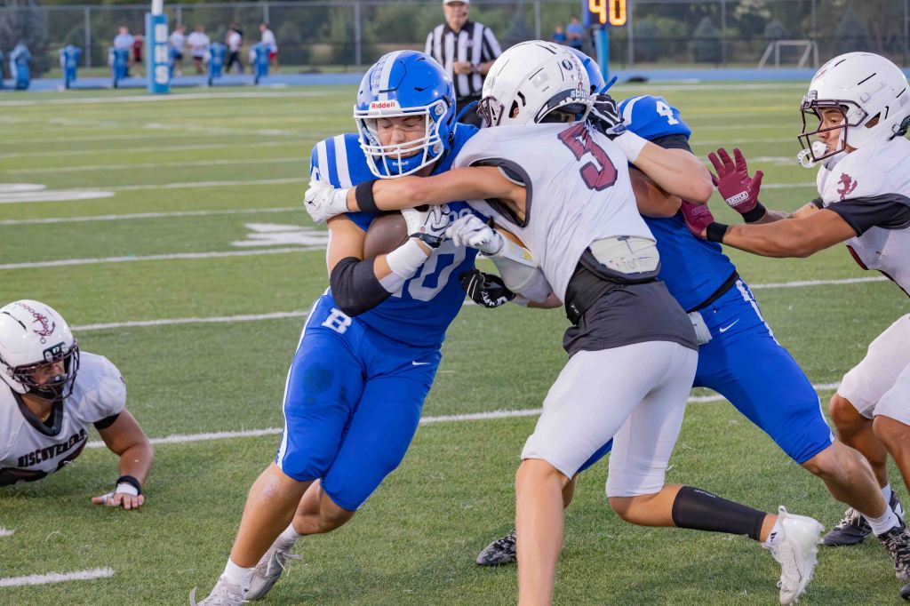 A football player in a blue jersey is being tackled by two opponents in white jerseys during a game, while another player lies on the field in the background.