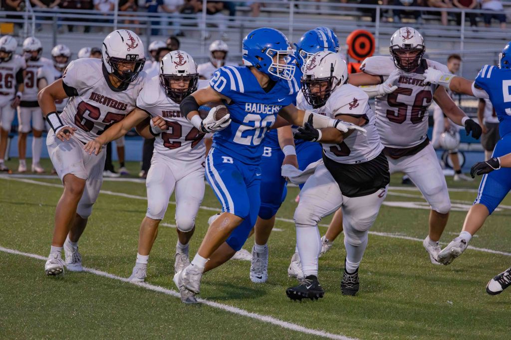 A high school football player in blue jersey number 20 evading multiple opponents in white jerseys during a game on a football field.
