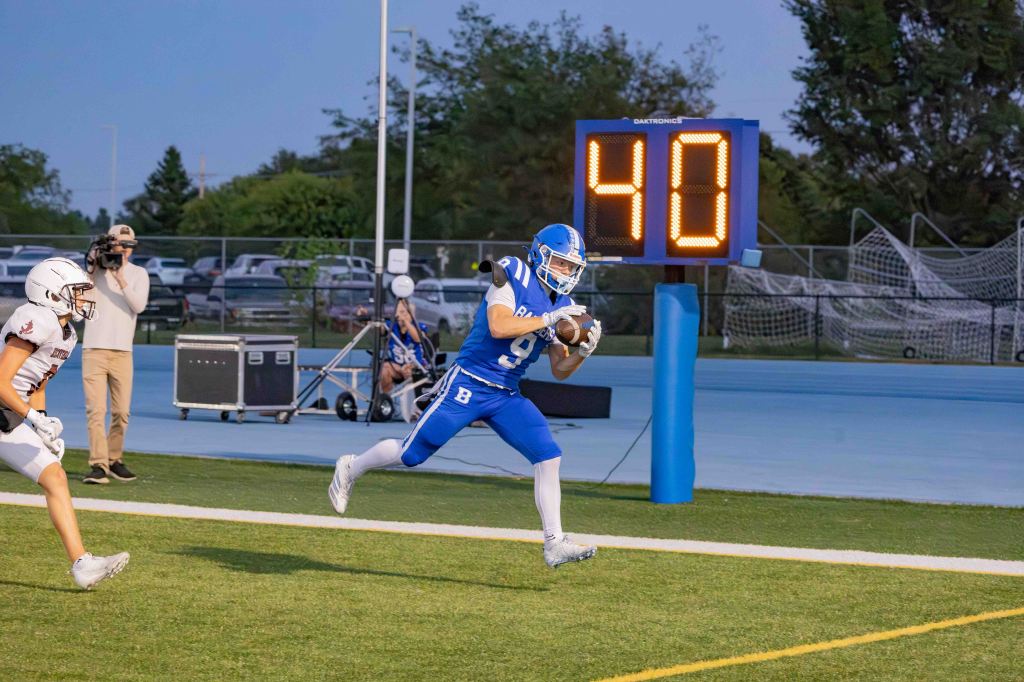 A football player in blue uniform running for a touchdown, with a digital scoreboard displaying the number 40 in the background, while an opposing player in a white uniform trails behind.