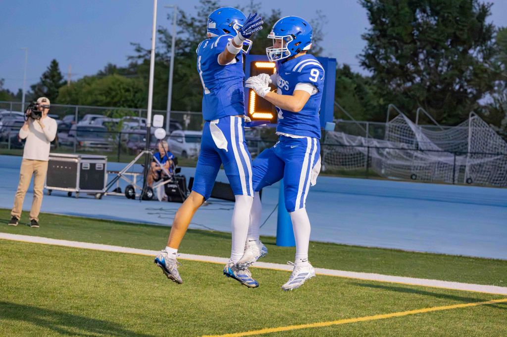 Two football players in blue uniforms celebrating a touchdown by jumping and high-fiving on the field, with a camera and spectators in the background.