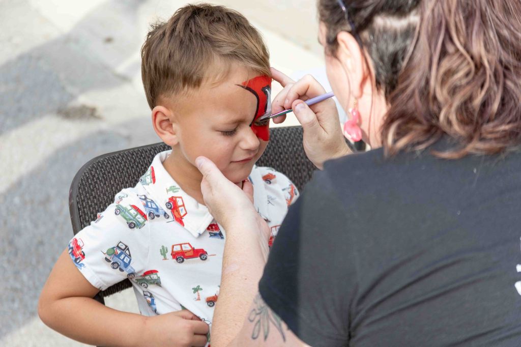 A child sitting in a chair having their face painted, with a woman applying red paint on the child's forehead.