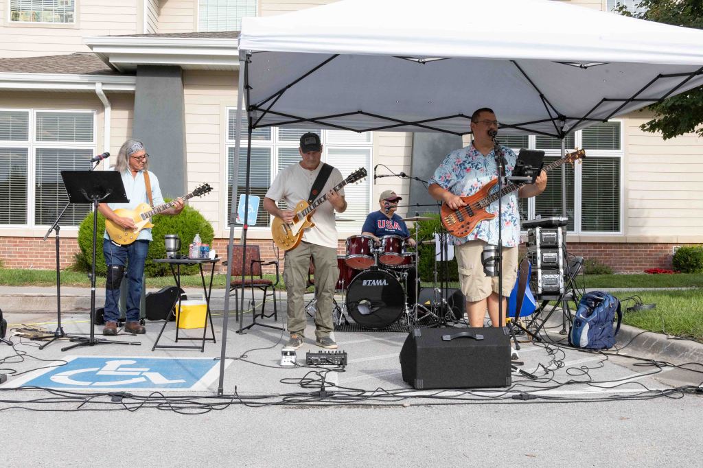 A four-member band performing outdoors under a tent. The musicians include two guitarists, a drummer, and a bassist. They are playing near a building, with instruments and equipment set up on the ground.