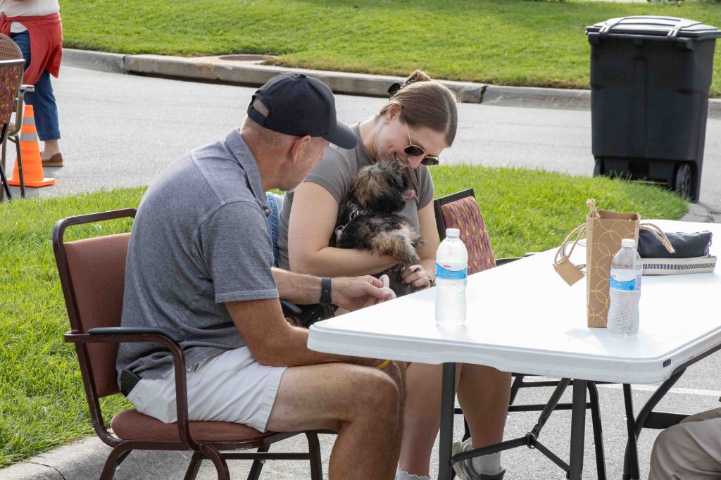 A woman holds a small dog while sitting at a table with a man, both engaged in conversation. Nearby, there are water bottles and a bag on the table, with a grassy area and a trash bin in the background.