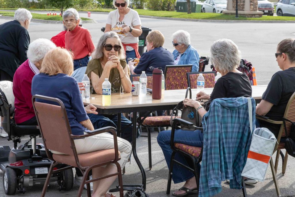 A group of elderly women gathered around a table outdoors, some seated and others standing, engaging in conversation and enjoying refreshments on a sunny day.