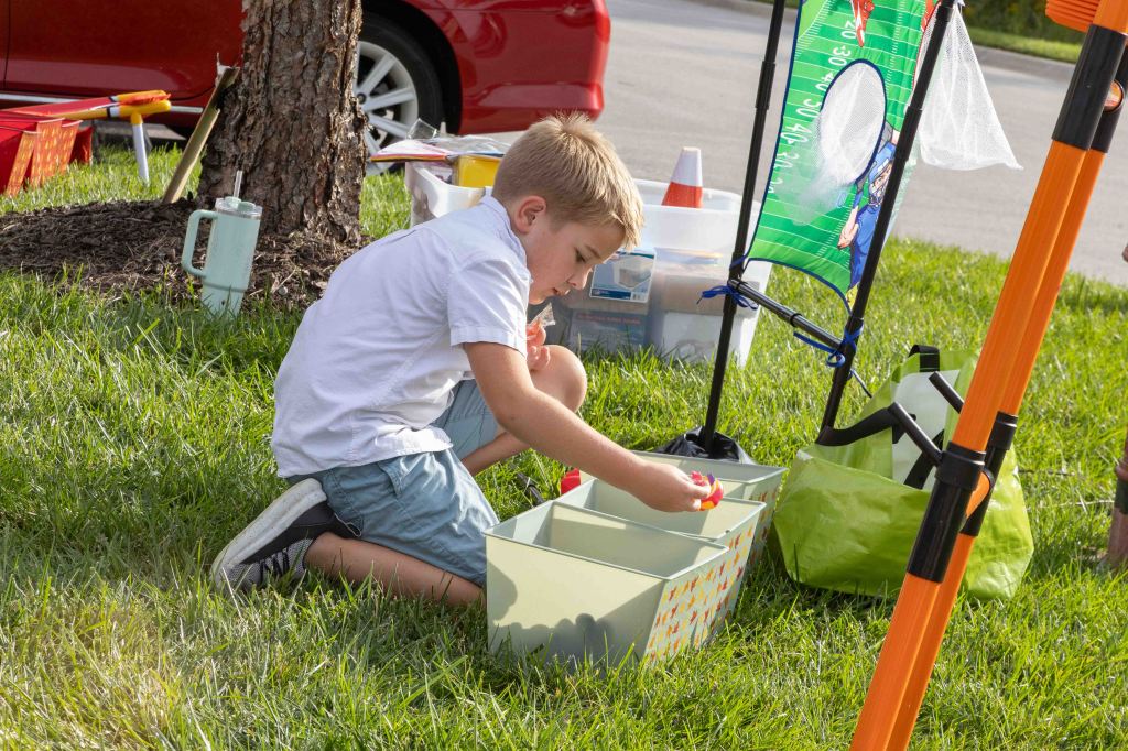 A young child kneeling on grassy ground, sorting colorful items into storage boxes. In the background, there are various outdoor play equipment and a red vehicle.