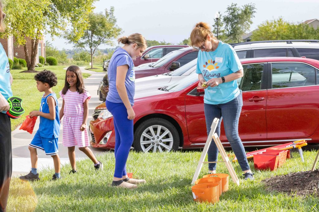 A group of children and adults participate in a community event outdoors. Two children are walking along a pathway, while an adult in a blue shirt engages with activities using orange buckets and a wooden stand in front of parked cars.