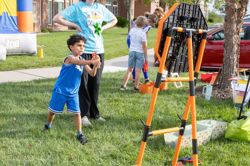 A young boy in a blue outfit is throwing an object at a colorful game target, while a woman and other children are engaged in activities in the background.