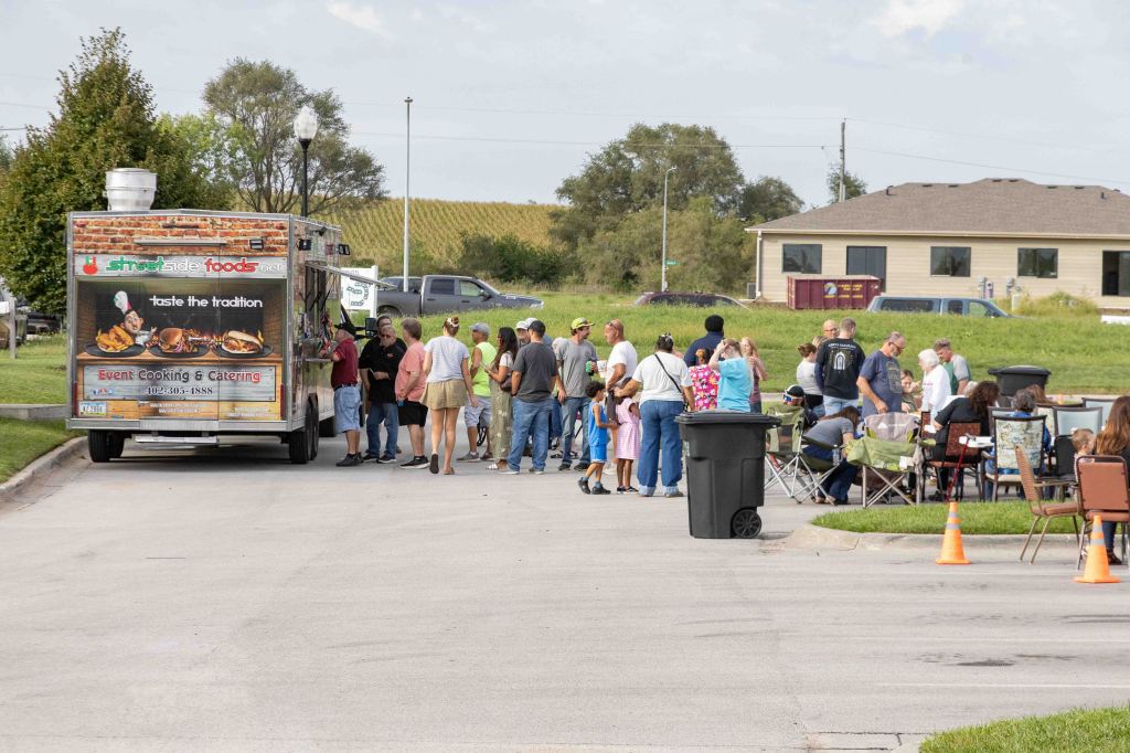 A food truck with the branding 'Street Side Foods' parked on a street, with a crowd of people waiting in line. Several individuals are seated in chairs nearby, enjoying the outdoor gathering.