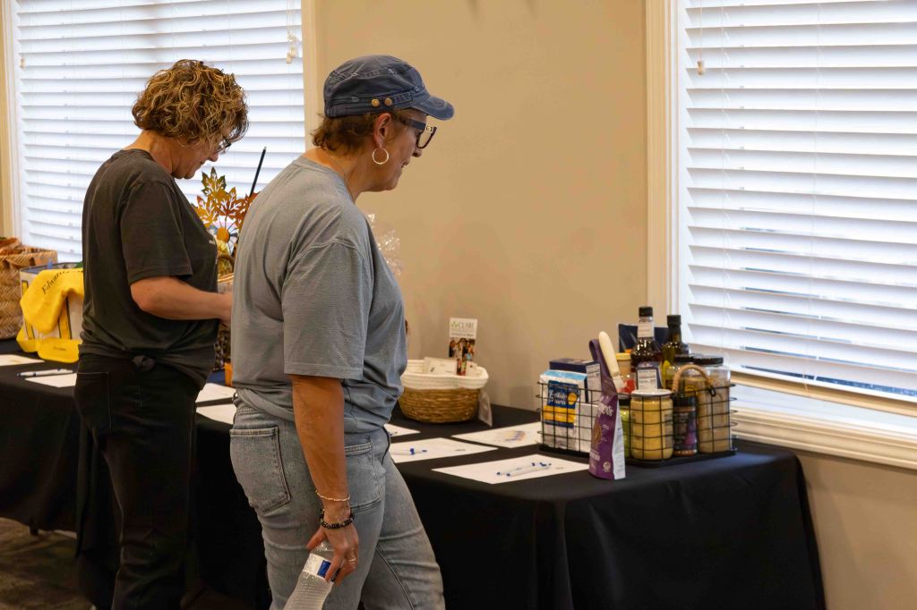 Two women are observing a display of items on a table, with one woman taking notes and the other looking closely at the offerings. The table features various products, including food items, a basket, and printed materials, set against a backdrop of a well-lit room with window blinds.