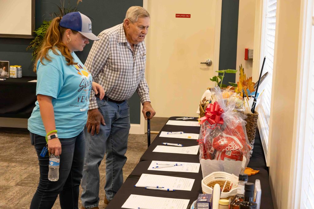 A woman and an older man examine items on a table at an event, with various gift baskets and notes visible.