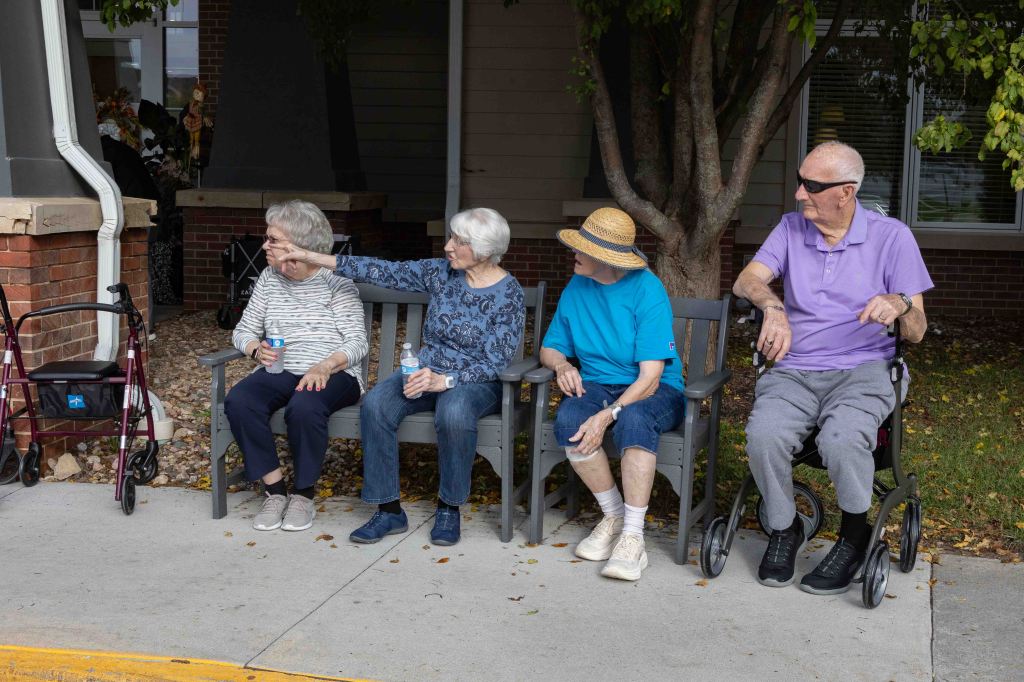 Four elderly individuals sitting on benches outdoors, sharing drinks and smiling. They are positioned next to a tree and a building, with a walker in the foreground.