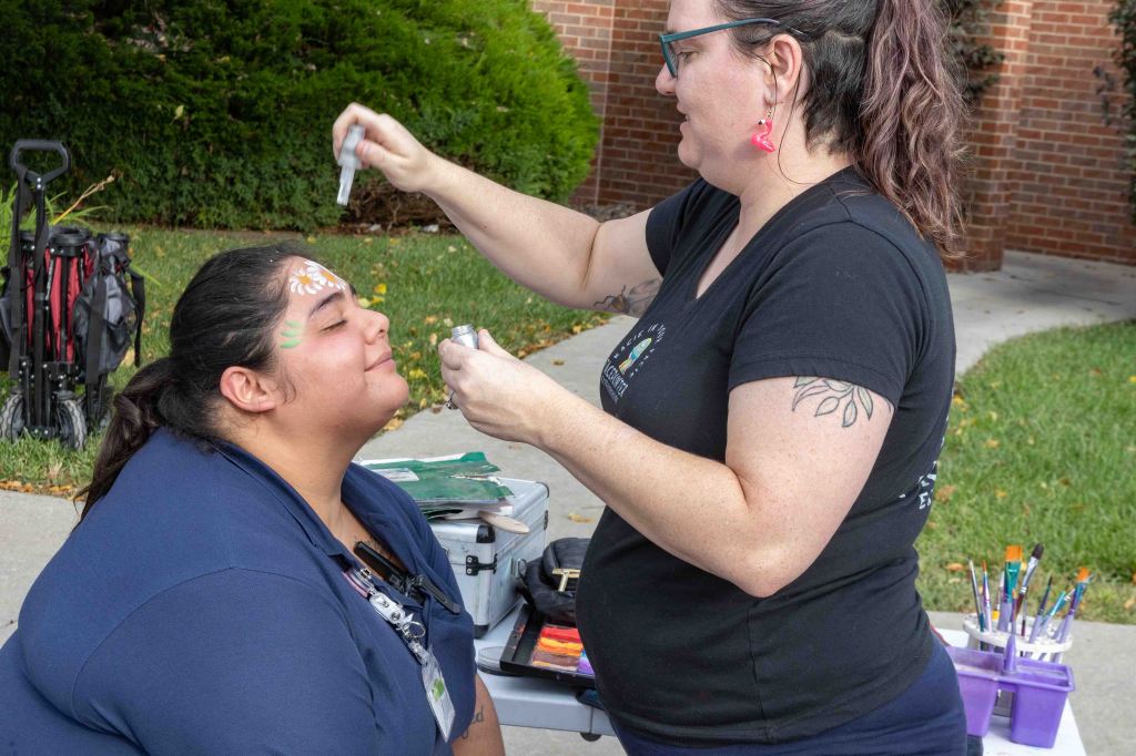 A woman is applying face paint to another person outdoors, with face painting supplies visible in the background.
