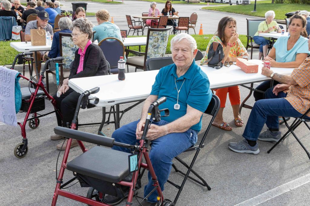 A senior man sitting at a table with a walker, surrounded by other elderly individuals enjoying a community event outdoors.