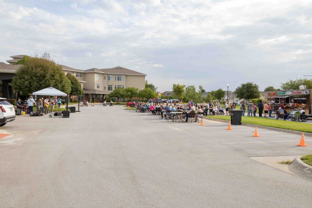 An outdoor gathering with people seated at tables and a food truck in a residential area, featuring a stage with musicians and a festive atmosphere.