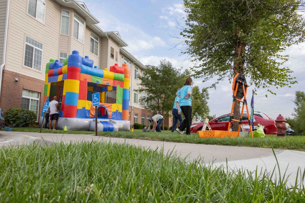 An inflatable bounce house in a residential area with people setting up nearby. The scene includes a tree, some landscaping, and a parked car.