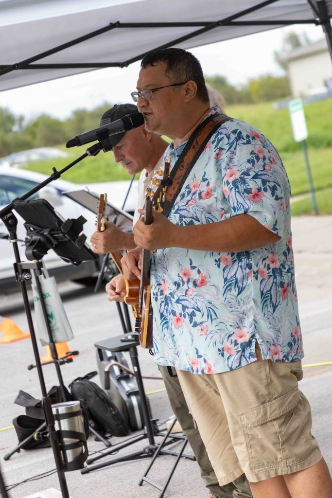 A musician in a floral shirt plays guitar while singing into a microphone, with another musician partially visible in the background.